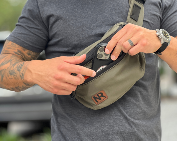 The OG Nut Ruck in Ranger Green being worn cross body on an athletic built man with tattoos. he is wearing a watch with a black band and is placing a cellular smart phone device in the front zipper pocket of the packet.