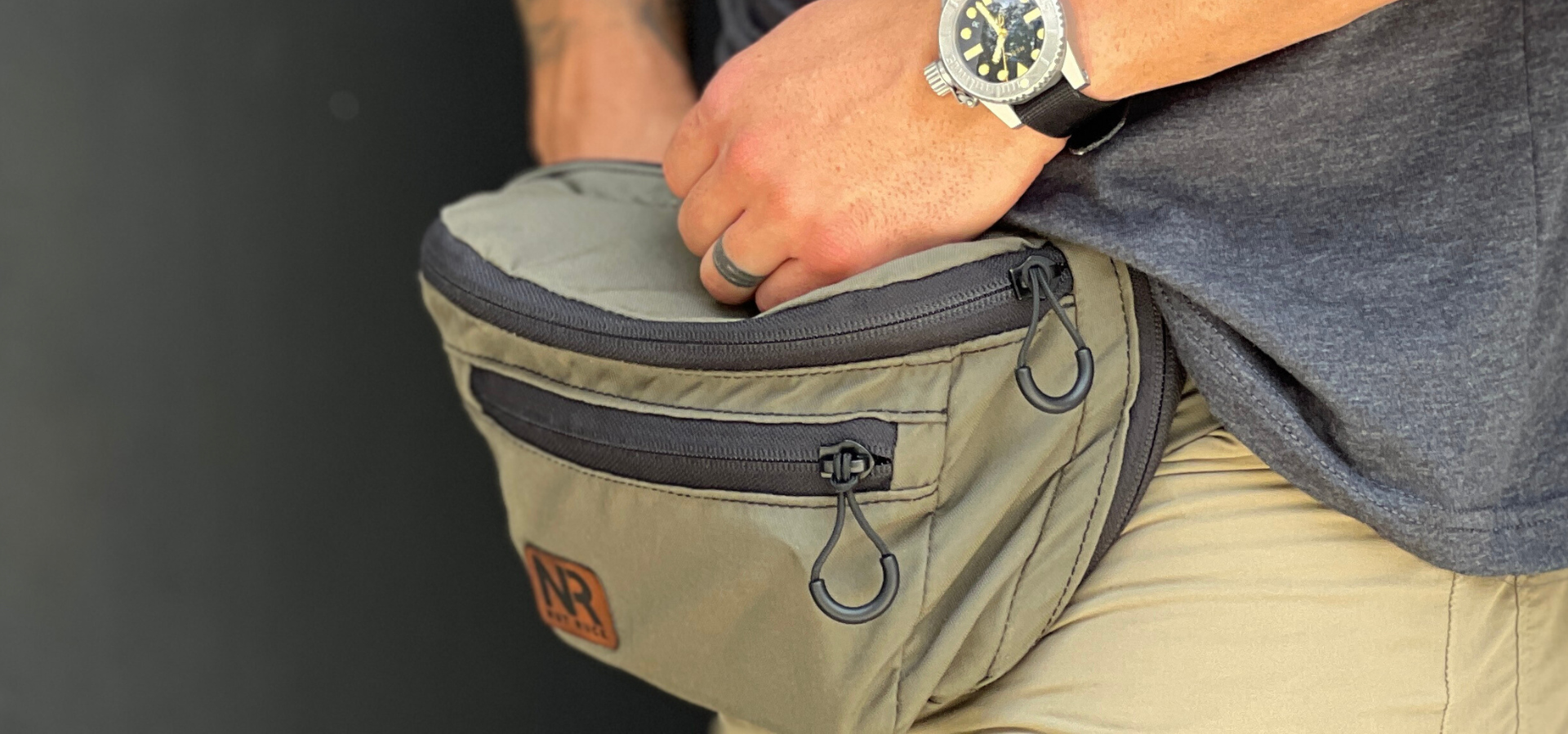 The OG Nut Ruck waist pack in Ranger Green with black zippers on a man with a dark heather grey shirt and tan shorts with a watch and wedding band.